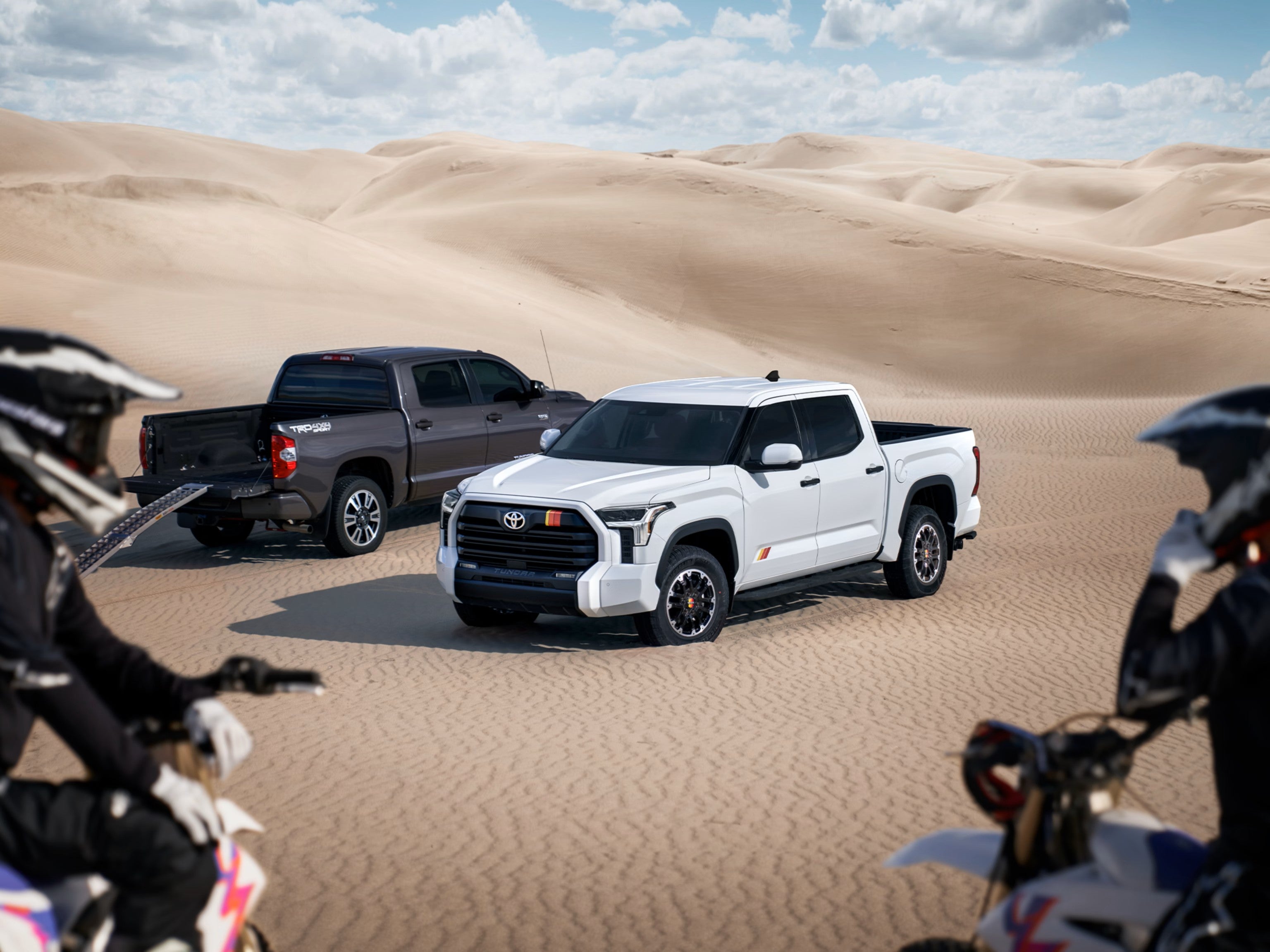 White Toyota Tundra with the TRD Rally Package on a sand dune with a dark Tundra behind it and two dirt bike riders in front